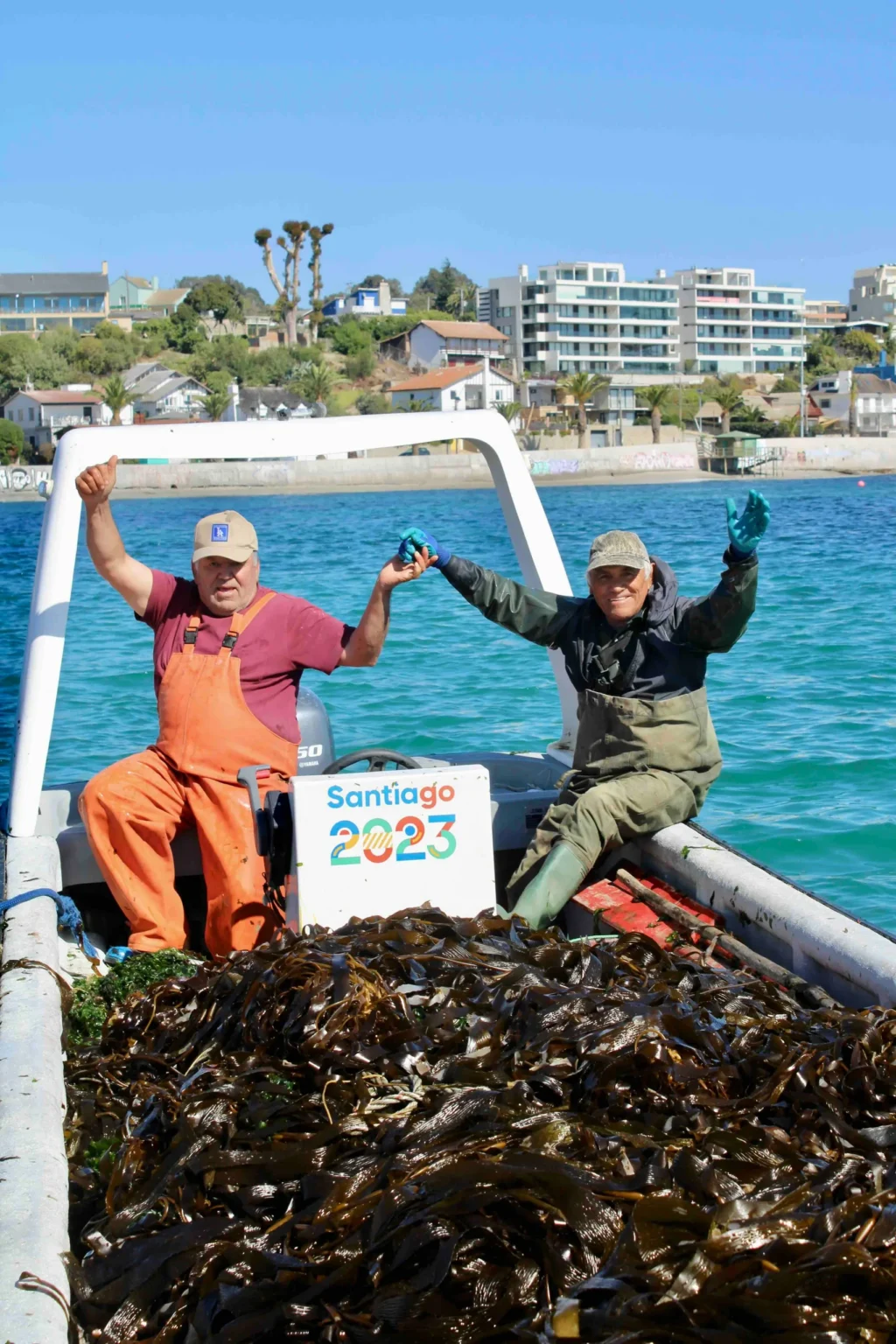Dos pescadores celebran en una lancha cargada de algas frente a la costa